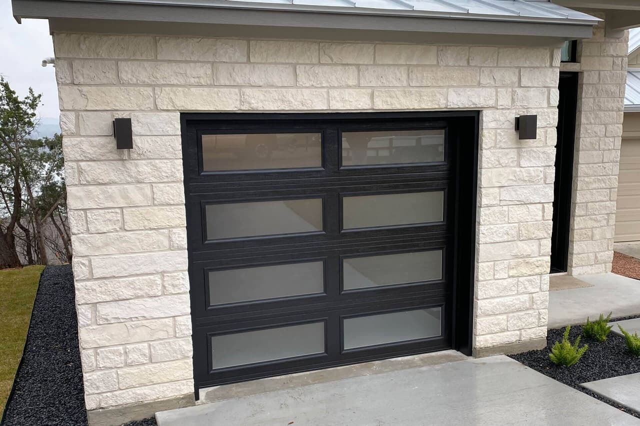 Modern black steel garage door with frosted glass panels installed by Door Pro Plus on a stone home in Marble Falls, TX