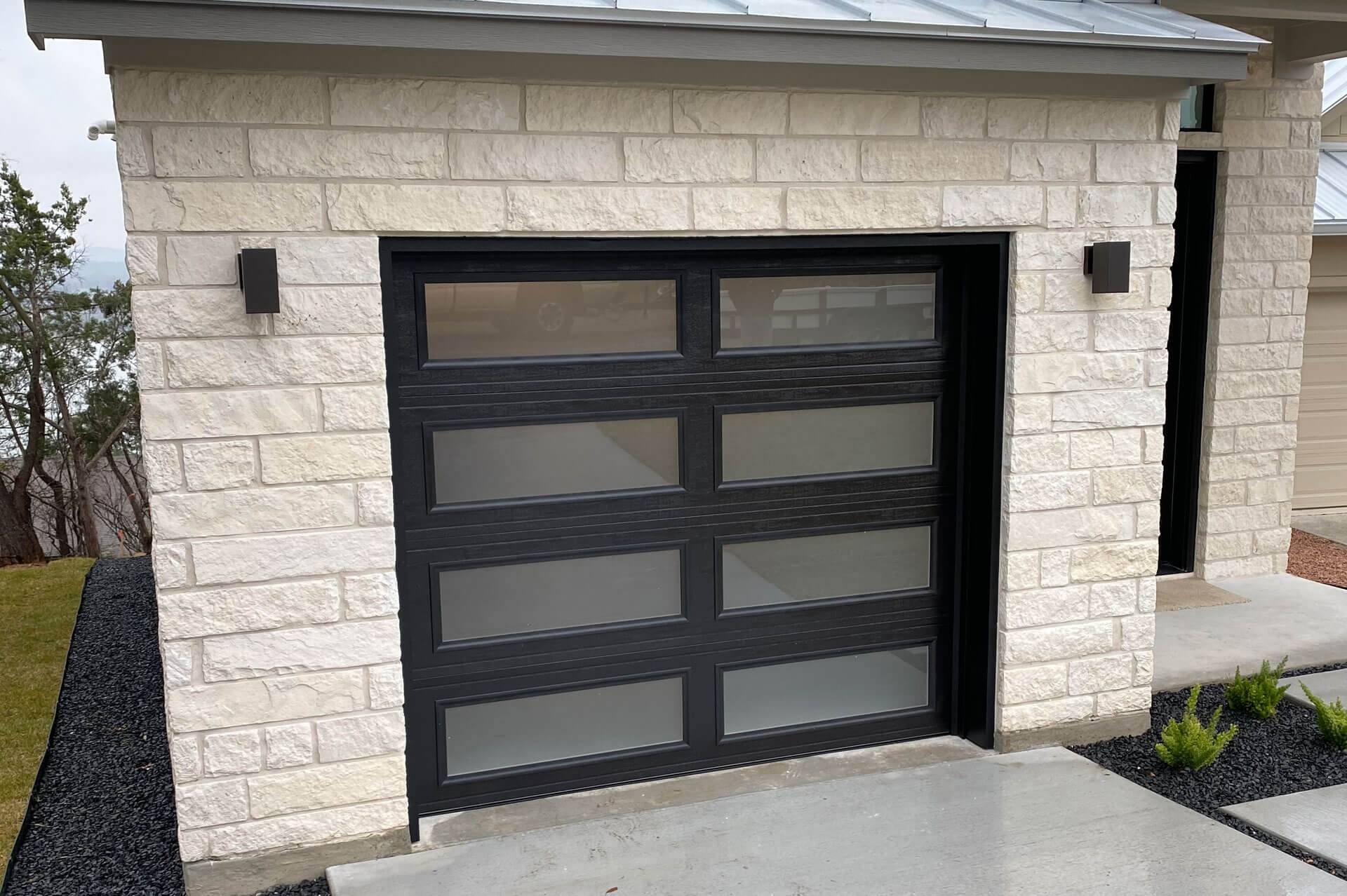 Modern black steel garage door with frosted glass panels installed by Door Pro Plus on a stone Hill Country home
